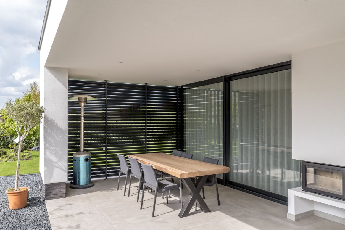 Terrasse moderne avec table en bois, chaises grises, chauffage extérieur et murs en verre, entourée de verdure.