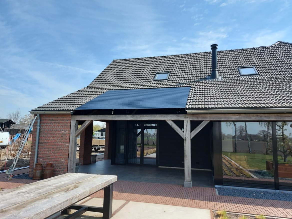 Maison moderne avec toiture en tuiles, panneaux solaires et large terrasse, entourée de verdure sous un ciel dégagé.