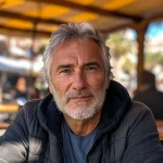 Homme au regard intense, cheveux gris, barbu, assis à une terrasse ensoleillée, ambiance détendue et conviviale. {JPEG}