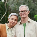 Un couple senior souriant, entouré de verdure, se tient près l'un de l'autre dans un cadre naturel apaisant. {JPEG}