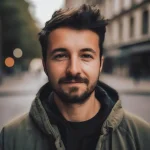 Portrait d'un jeune homme souriant, avec une barbe et des cheveux bruns, en veste, devant un décor urbain flou. {JPEG}