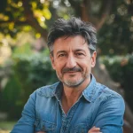 Homme souriant aux cheveux gris, portant une chemise en denim, se tenant dans un jardin verdoyant. {JPEG}