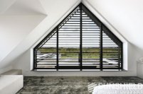 Fenêtre triangulaire avec volets noirs, vue sur un paysage extérieur, pièce moderne avec moquette et mobilier élégant.